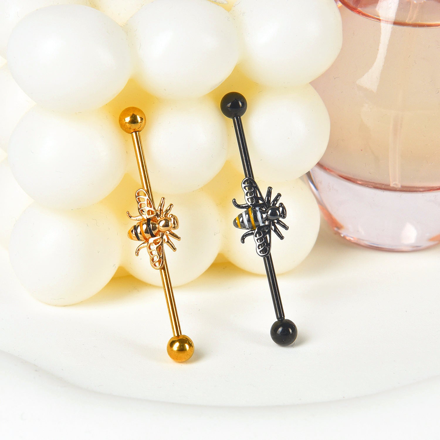 Two industrial ear piercing barbells with bee centerpieces standing vertically against a decorative white bubble candle