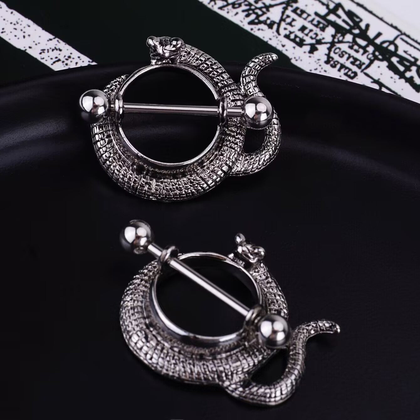 Two snake-shaped nipple shields on a black tray with a gothic script background